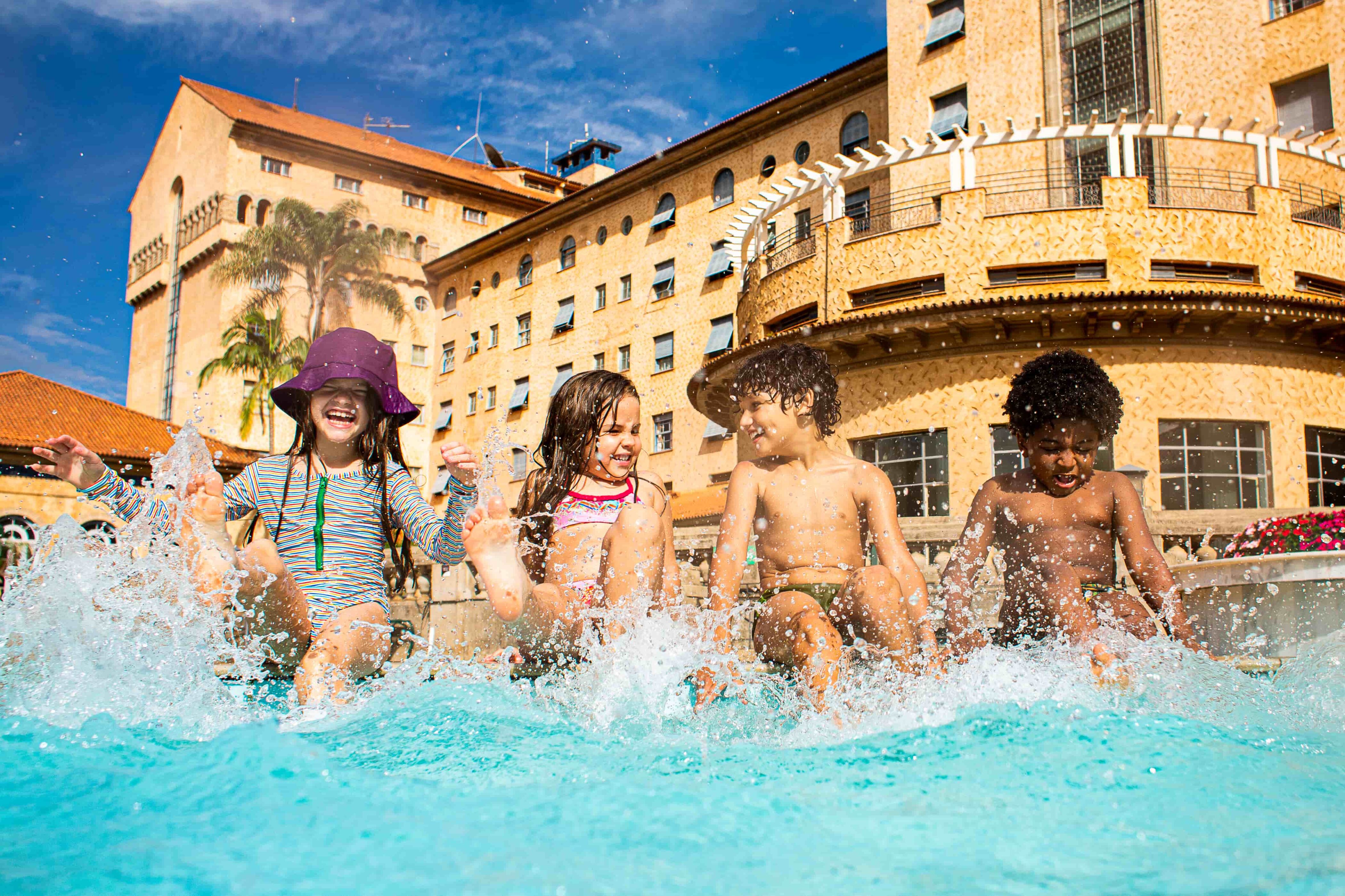 crianças brincando na piscina em frente à fachada do Grande Hotel, sentadas na borda, batendo os pés na água.